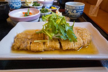 Hafif sosta servis edilmiş, dikdörtgen bir tabakta taze yapraklı yeşilliklerle süslenmiş gevrek domuz pirzolası. Çeşitli mezeler ve seramik kaplar samimi, davetkar bir yemek sahnesi yaratır..