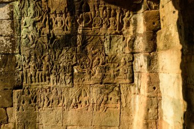 Ayrıntılı kumtaşı yardım panelleri Angkor, Kamboçya 'daki Bayon tapınağının antik duvarlarında tarihi ve mitolojik sahneleri tasvir eder. Sıcak güneş ışığı, Khmer işçiliğinin sanatını ve derinliğini ortaya çıkaran desenli oymaları vurguluyor..