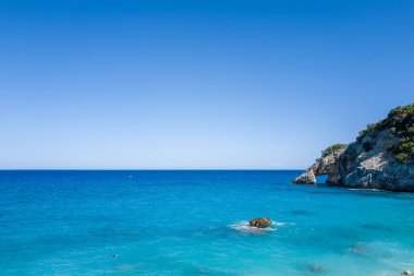 Canlı turkuaz sular, Cala Goloritze, Baunei, Sardinya 'da doğal bir kemerle dramatik bir kireçtaşı uçurumuna çarpar. Sahne açık mavi bir gökyüzü altında güneş ışığıyla yıkanmış, yaz sükuneti hissi uyandırıyor..