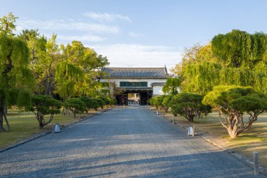 Yeşil ağaçlarla çevrili düz çakıl yolu Japonya 'nın Kyoto kentindeki Nijo Kalesi' nin kiremitli çatısına sahip geleneksel ahşap bir kapıya çıkar..