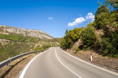 Sardinya 'nın Baunei ve Dorgali yakınlarında canlı mavi bir gökyüzünün altında yemyeşil tepeler ve kayalık yamaçlarla çevrili asfalt yol. Manzara güneş ışığıdır, açıktır ve doğal dokular ve canlı yapraklarla sükunettir..