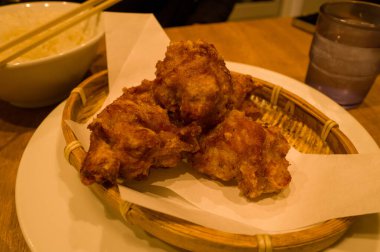 Japon usulü kızarmış tavuk parçaları, örülmüş bambu bir sepetin içinde, sıcak bir Osaka restoranında, ahşap bir masanın üzerinde bir kase pirinç ve bir bardak su ile parşömen kağıdı üzerinde servis edilir. Sıcak aydınlatma çıtır çıtır dokuyu ve davetkar rengi vurgular.