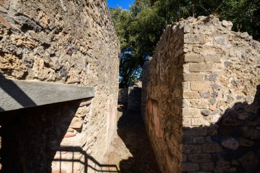 İtalya 'nın Pompeii kentindeki arkeolojik bölgede yıpranmış volkanik taş duvarlarla çevrili dar bir sokak. Benekli güneş ışığı, eski yapıların ötesindeki yeşilliklerle pürüzlü duvar dokularını ve derin gölgeleri vurguluyor..
