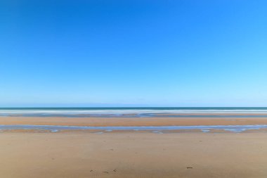 Uçsuz bucaksız kumlar ve sığ gelgit havuzları Omaha Sahili boyunca uzanır bulutsuz, canlı mavi gökyüzünün altında, huzurlu ve geniş bir kıyı manzarası yaratır..