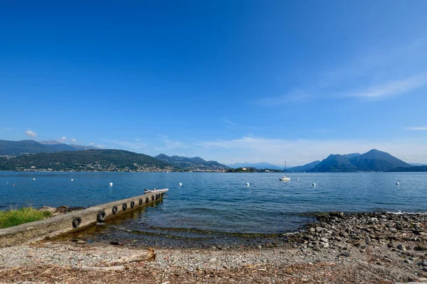 Maggiore Gölü 'nün berrak mavi sularına uzanan taştan bir iskelenin olduğu sakin bir göl manzarası dağınık şamandıralar ve ıssız bir yelkenli. Rocky kıyı şeridi ve uzak orman tepeleri kuzeydeki bulutsuz gökyüzünün altında barışçıl bir zemin oluşturur.