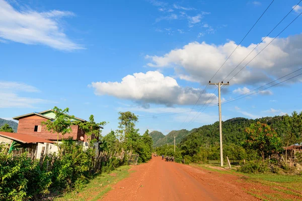 Güneşli kırmızı toprak yol ahşap evleri ve yeşil bitki örtüsünü geçerek parlak mavi gökyüzünün altındaki uzak tepelere doğru uzanıyor. Güç hatları yol boyunca uzanıyor. Dağınık bulutlar ve sakin kırsal atmosferle..