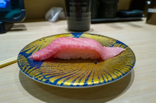 Bir parça yağlı ton balığı nigiri soluk ahşap tezgahın üzerinde çarpıcı mavi ve altın rengi bir güneş patlaması tabakası üzerinde sunuluyor. Mermer pembe balıklar ve parlak pirinçler modern bir Japon restoranındaki yumuşak kapalı mekan ışıklarıyla aydınlatılır..