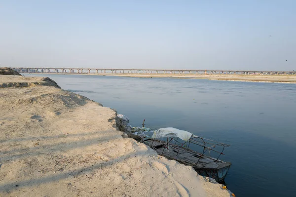 Basit bir çerçeve ile yıpranmış ahşap bir tekne, Hindistan 'ın Prayagraj kentindeki Ganj Nehri' nin kumlu kıyısında yer alır. Sakin su ve berrak bir gökyüzünün altındaki uzak köprü sakin ve geniş bir atmosfer yaratır..