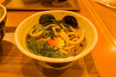 Bir seramik kase udon noodle çorbası kalın erişte, dilimlenmiş yeşil soğan, deniz yosunu ve şeffaf et suyunda dekoratif sebze dilimi içerir. Ilık ışıklandırma, parlak dokuyu ve ahşap tepside sunumu vurgular.