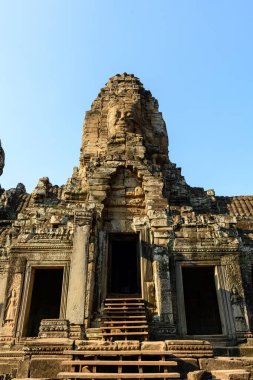 Ilık güneş ışığı, Angkor, Kamboçya 'daki Bayon Tapınağı' nın ana girişinde yükselen yıpranmış kumtaşı yüz kulesini vurguluyor. Karmaşık Khmer oymaları ve antik mimari ayrıntılar açık mavi bir gökyüzü tarafından çerçeveleniyor ve anıtlara vurgu yapılıyor
