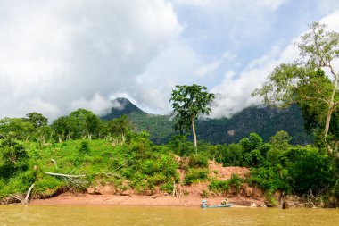 Kuzey Laos 'taki Muang Ngoi yakınlarındaki Nam Ou nehri boyunca yemyeşil bir nehir kıyısında alçak bulutlar sürükleniyor. Yoğun bitki örtüsü ve aşınmış toprak sahneyi oluştururken, küçük bir tekne sakin nehir manzarasına ölçek ekliyor..