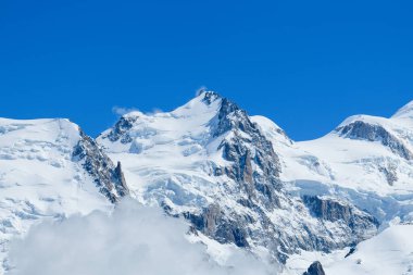 Mont Maudit 'in engebeli, karla örtülü zirvesi sürüklenen bulutların üzerinde yükselir. Pürüzlü kaya yüzeyleri ve Fransız Alpleri' nin derin mavi gökyüzünün altında keskin bir şekilde tanımlanan buzlu yamaçlar..