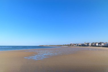 Geniş ıslak kumlar sakin denize doğru uzanır, Grandcamp-Maisy köyü bulutsuz mavi gökyüzünün altında uzak kıyıları kaplar. Sahne açık, parlak ve huzurlu, kumsaldaki ince yansımalarla.