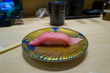 Bir parça yağlı ton balığı nigiri, Japon restoranındaki soluk ahşap bir tezgahın üzerinde çarpıcı mavi ve sarı desenli bir tabağın üzerinde oturur. Mermer pembe balıklar ve parlak pirinçler, yumuşak iç mekan ışıklarıyla aydınlatılarak taze ve davetkar bir ortam yaratıyorlar.