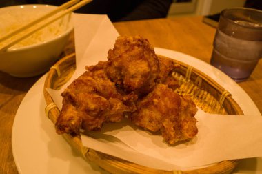 Kızarmış tavuk kareage, bir kase pirinç ve bir bardak su eşliğinde, ahşap bir masanın üzerindeki parşömenle örülmüş bir bambu sepetinde servis edilir. Sıcak aydınlatma, yemeğin çıtır çıtır dokusunu ve altın rengini vurgular..