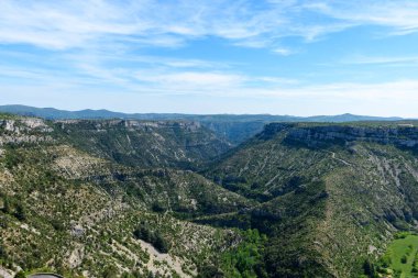 Navacelles Sirki 'nin engebeli kireçtaşı uçurumları ve parlak mavi gökyüzünün altındaki sarp yeşil yamaçlar geniş bir manzara. Güneş ışığı, dramatik hatları ve kanyonun vahşi doğal dokusunu vurgulayan canlı gölgeler oluşturur..