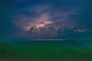 Karanlık bir dağ manzarasının üzerinde gece vakti yükselen fırtına bulutları arasında uzak bir şimşek çakar. Sahnede dramatik bulut oluşumları, katmanlı siluetler ve kasvetli bir alacakaranlık gökyüzü yer alıyor..