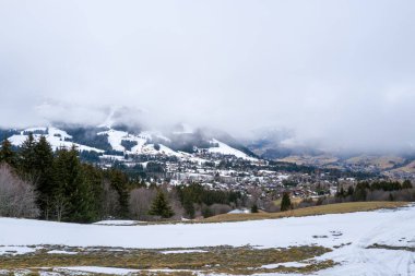 Megeve köyünün panoramik manzarası karlı bir dağ vadisinde, eriyen karlar ve alçak asılı bulutlardan oluşan kalın bir battaniyenin altında ebedi yeşil ağaçlar. Karamsar kış atmosferi beyaz yamaçlar ve açığa çıkan yamaçlar arasındaki zıtlığı vurgular.