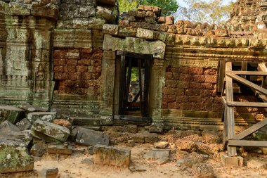 Karmaşık oymaları olan antik taş kapı, Kamboçya Angkor bölgesindeki Ta Som Tapınağı 'nda yıpranmış bir kumtaşı duvarın içinde duruyor. Yıkılmış duvarcılık ve ahşap destek sıcak öğleden sonra ışığı ve doğal dokular arasında tarihi yapıyı vurguluyor.