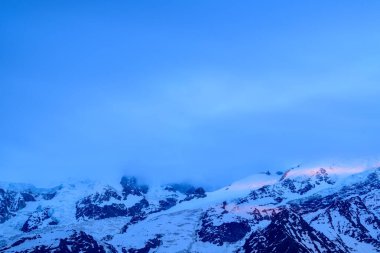 Mont Blanc 'ın buzlu zirveleri ve etrafındaki dağlar serin mavi ışıkla yıkanır. Güneşin doğuşundan gelen yumuşak pembe ipuçları karları ve sakin bir gökyüzünün altındaki buzulları aydınlatır..