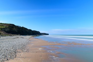 Pürüzsüz dalgalar Omaha Sahili 'nin kumlu ve çakıl taşlı sahillerini silip süpürüyor, engebeli uçurumlar kıyı şeridi boyunca canlı mavi bir gökyüzünün altında yükseliyor. Sahne sakin, geniş ve doğal görünüyor..