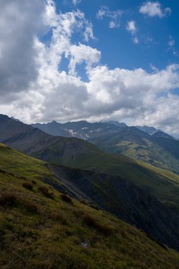 Val Ferret 'in dik çimenli tepeleri gölgeli tepelere doğru iniyor, dokulu bulutlar engebeli dağ manzarasının üzerinde canlı mavi bir gökyüzünde sürükleniyor..