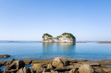 Japonya 'nın Shirahama kıyısındaki Iconic Engetsu Adası, parlak, bulutsuz bir gökyüzü altında, ön planda engebeli kayalar ve doğal kemerin üzerindeki yemyeşil yemyeşil yeşil sulardan yükselir..