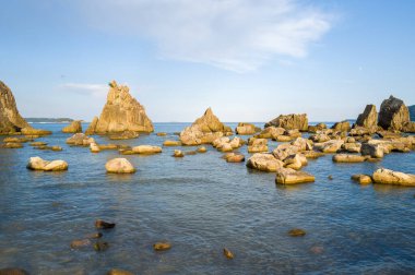 Hashiguiwa, Kushimoto, Japonya 'daki sakin mavi denizden yükselen altın taş çıkıntıları ve dağınık kayalar, açık bir gökyüzü altında sıcak öğleden sonra ışığı ile aydınlatılıyor..