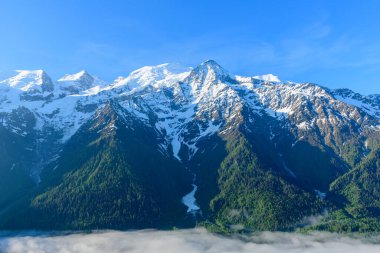Parlak sabah güneşi, Mont Blanc kalabalığının dramatik karlı zirvelerini aydınlatıyor. Alt yamaçları kaplayan koyu yeşil ormanlar ve üste sürüklenen sis zerreleri..
