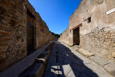 İtalya 'nın Pompeii arkeoloji sahasında volkanik taş ve tuğla duvarlarla çevrili yıpranmış bir Roma karayoluna uzun bir bakış açısı. Sert asfalt ve tarihi dokular berrak mavi gökyüzünün altında güçlü güneş ışığıyla aydınlatılır..