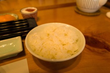 A bowl of freshly steamed white rice is placed on a polished wooden table, surrounded by elegant Japanese tableware and warm ambient lighting, creating a cozy morning atmosphere.