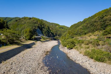 Japonya 'nın Wakayama kentindeki Kumano Kodo nehri boyunca açık mavi bir gökyüzünün altında yemyeşil, ormanlık tepeler arasında sığ bir nehir akmaktadır. Güneş ışığı taşları ve yeşilliği aydınlatır, sakin bir kırsal alan yaratır..