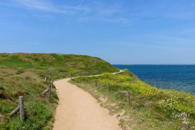 Ahşap direklerle ve çiçek açan kır çiçekleriyle dolu kıvrımlı patika Saint Pierre Quiberon 'da Pointe Beg-An-Aud' daki çimenli burnu takip ediyor, berrak bir gökyüzünün altındaki derin mavi denize bakıyor..
