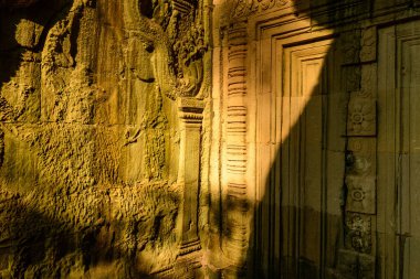 Sıcak güneş ışığı, Kamboçya Angkor bölgesindeki Ta Prohm tapınağının karmaşık kumtaşı duvarı ve kapı aralığında dramatik gölgeler oluşturur. Yıpranmış yüzey ayrıntılı Khmer motiflerini ve doğal dokularını ortaya çıkararak çarpıcı bir etkileşim yaratıyor.