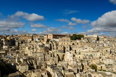 Matera, İtalya 'nın geniş bir manzarası, sergiler sıkıca toplanmış kireçtaşı evler ve güneş ışığıyla aydınlatılmış antik mimari tüylü bulutlarla kaplı canlı mavi gökyüzü.