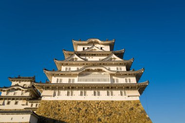 Himeji Kalesi 'nin ön cephe manzarası katmanlı beyaz duvarlar ve süslü kiremitlerle döşenmiş taş bir temelin üzerinde yükselen, canlı, bulutsuz mavi gökyüzüne karşı kurulmuş çatılar..