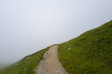 Issız bir toprak yol, çimli bir yamaç boyunca süzülür, yavaş yavaş kalın beyaz sise dönüşür ve dağ manzarasını sarar ve sessiz, gizemli bir sahne yaratır..