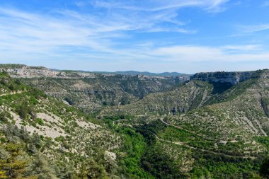 Navacelles Sirki 'nin engebeli kireçtaşı uçurumları, dolambaçlı yolları ve parlak mavi gökyüzünün altındaki yemyeşil bitki örtüsünün geniş manzarası. Dramatik arazi ve katmanlı dokular uçsuz bucaksız ve doğal bir güzellik hissi yaratır..