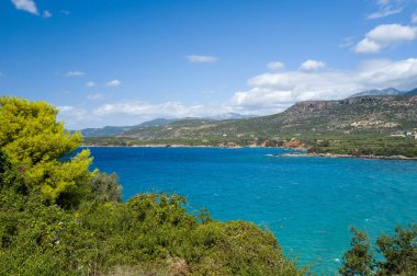 Parlak turkuaz su kıyı şeridi boyunca yoğun yeşil bitki örtüsüyle buluşur, engebeli tepeler ve dağınık bulutlu güneşli bir gökyüzünün altında dağınık evler. Manzara canlı ve taze, canlı bir Akdeniz yazını anımsatıyor..