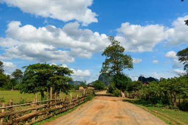 Kırmızımsı bir toprak yolu ahşap çitler ve dağınık ağaçlar arasında büyük beyaz bulutlarla dolu bir gökyüzünün altındaki uzak tepelere doğru esiyor. Geleneksel kırsal evler ve yemyeşil alanlar, Güneydoğu Asya 'nın barışçıl bir kırsalını çağrıştırıyor