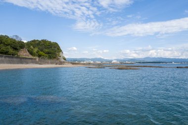 Japonya 'nın Tanabe kenti yakınlarında ufukta dağların göründüğü, parlak bir gökyüzünün altında kayalık bir kıyı şeridi ve yemyeşil yamaçla çevrili, geniş bir mavi deniz manzarası..