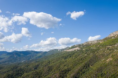 Sardunya 'nın Baunei yakınlarındaki dağınık beyaz bulutlarla kaplı parlak mavi bir gökyüzünün altında yemyeşil örtülü engin yuvarlanan tepeler. Güneş ışığı engebeli Akdeniz manzarasının dokusunu güçlendirir.