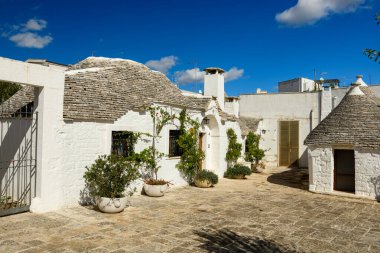 Apulia, İtalya 'da Alberobello' da dokulu taş çatılı geleneksel trullo evleriyle çevrili parlak bir taş avlu vardır. Akdeniz saksı bitkileri ve açık mavi gökyüzü kendine özgü mimari ve kırsal cazibeyi ön plana çıkarıyor..