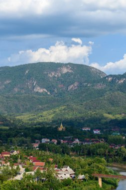 Luang Prabang, Laos 'un kıyısındaki yoğun tropikal ormandan geleneksel çatıları ve önlerinde bir nehri olan altın bir tapınak yükselir. Bu Güneydoğu Asya manzarasında yemyeşil tepeler ve dramatik bulutlar çarpıcı bir doğal zemin oluşturuyor..