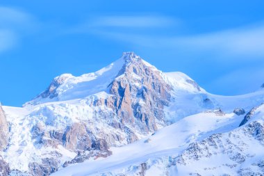 Mont Maudits 'in dramatik zirvesi buzlu yamaçların ve çıplak kayaların üzerinde yükseliyor, parlak güneş ışığıyla aydınlanıyor yumuşak bulutlu parlak gökyüzünün altında..