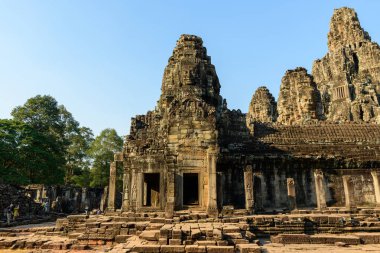 Angkor, Kamboçya 'daki Bayon Tapınağı' nın ana girişinde heybetli yüzlü antik taş kuleler yükseliyor. Yıpranmış kumtaşı ve karmaşık Khmer ayrıntıları sıcak öğleden sonra güneş ışığıyla aydınlatılır ve yemyeşil orman ağaçları tarihi çerçeveyi oluşturur.