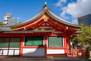 Japonya 'nın Kobe kentindeki bir Shinto türbesi pavyonunun canlı kırmızı ve beyaz cephesinde altın rengi detaylar ve yeşil kafes pencereleriyle zarif eğimli bir çatı bulunur. Güneş ışığı keskin gölgeler bırakıyor, mimari çizgileri şehrin arka planına vurguluyor.