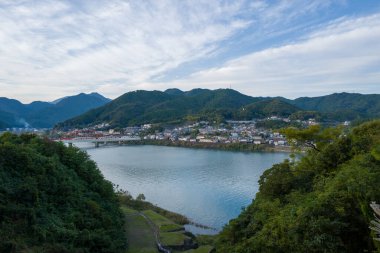 Huzurlu bir nehir kenarı, Japonya 'nın Shingu kentindeki geniş ve yumuşak bulutlu bir gökyüzünün altında sakin bir nehir kıvrılarak yemyeşil dağların eteklerinde yer alır..