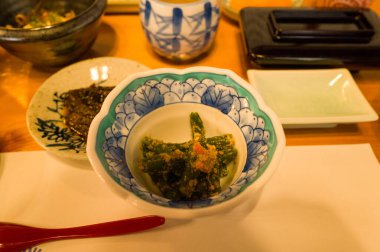 Süslü mavi ve beyaz bir porselen kasede susamlı yeşil fasulye servis edilir. Sıcak ışıklandırma geleneksel bir Japon kahvaltı ortamının dokularını ve renklerini vurgular.