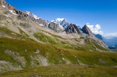 Canlı yeşil çayırlar ve kayalık çıkıntılar Col de la Seigne 'deki dramatik sivri tepelere doğru yükseliyor, açık mavi gökyüzünün altında Mont Blanc' ın karlı zirvesi. Güneş ışığı engebeli dokuları ve doğal hatları vurgular.